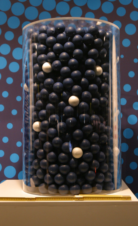 The colored image shows a cylinder-shaped vessel filled with many black and some white spheres. The vessel stands on a beige base and in front of a black wall with blue circles.