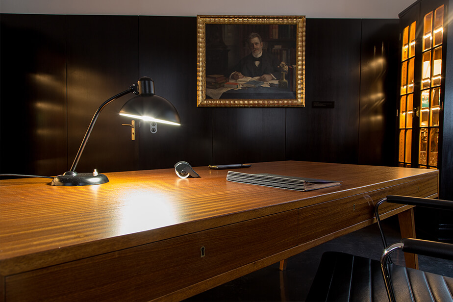 The photo shows the historical Rector’s Room at Universität Hamburg, replete with a fine wood desk, antique desk lamp, and a painting in the background.